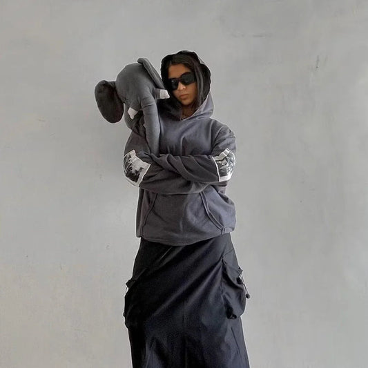 Person in dark streetwear hoodie and black skirt with elephant plush, urban minimalist style