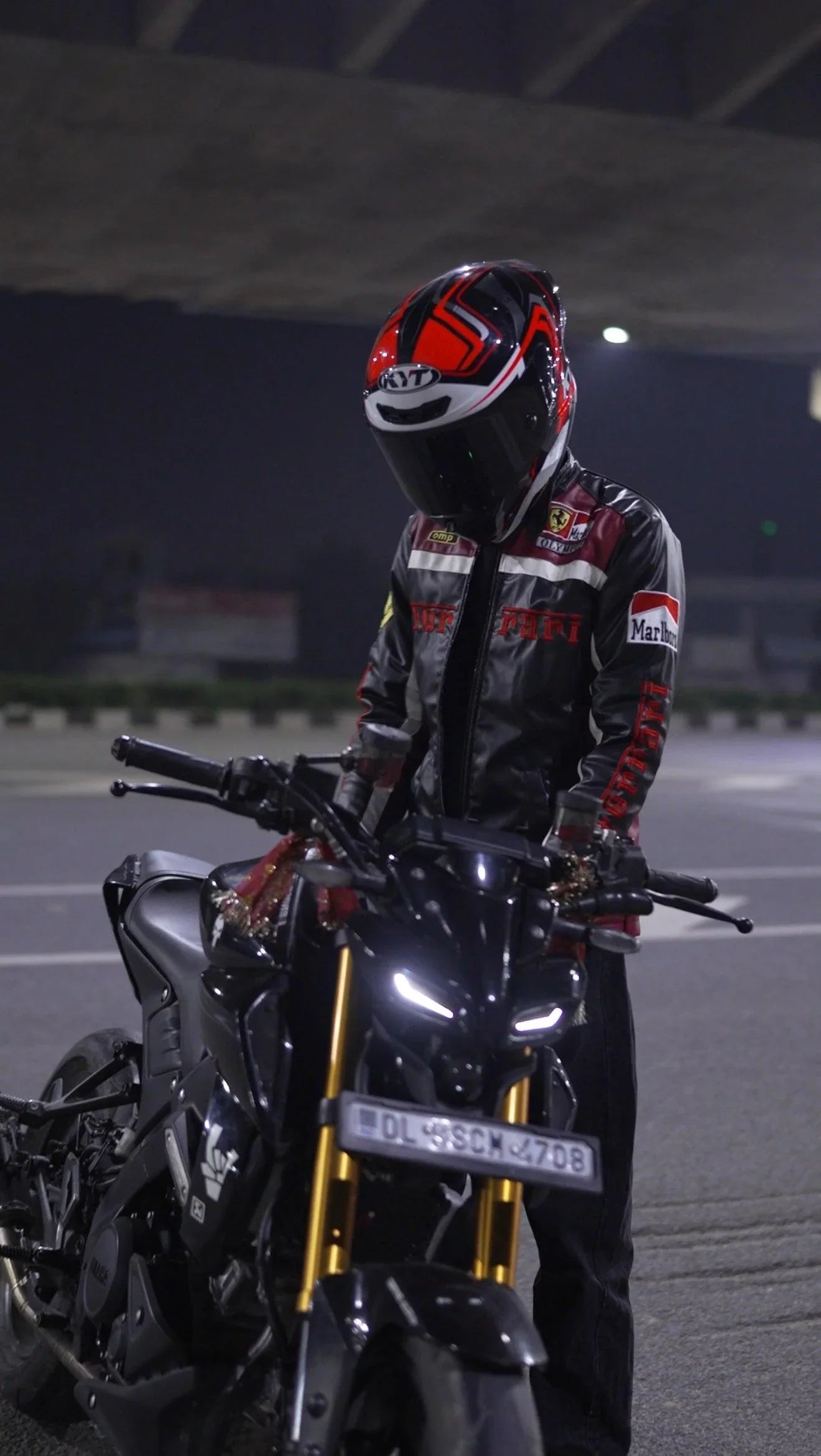 Man in a racing-inspired black leather jacket and helmet standing with a sport motorcycle at night
