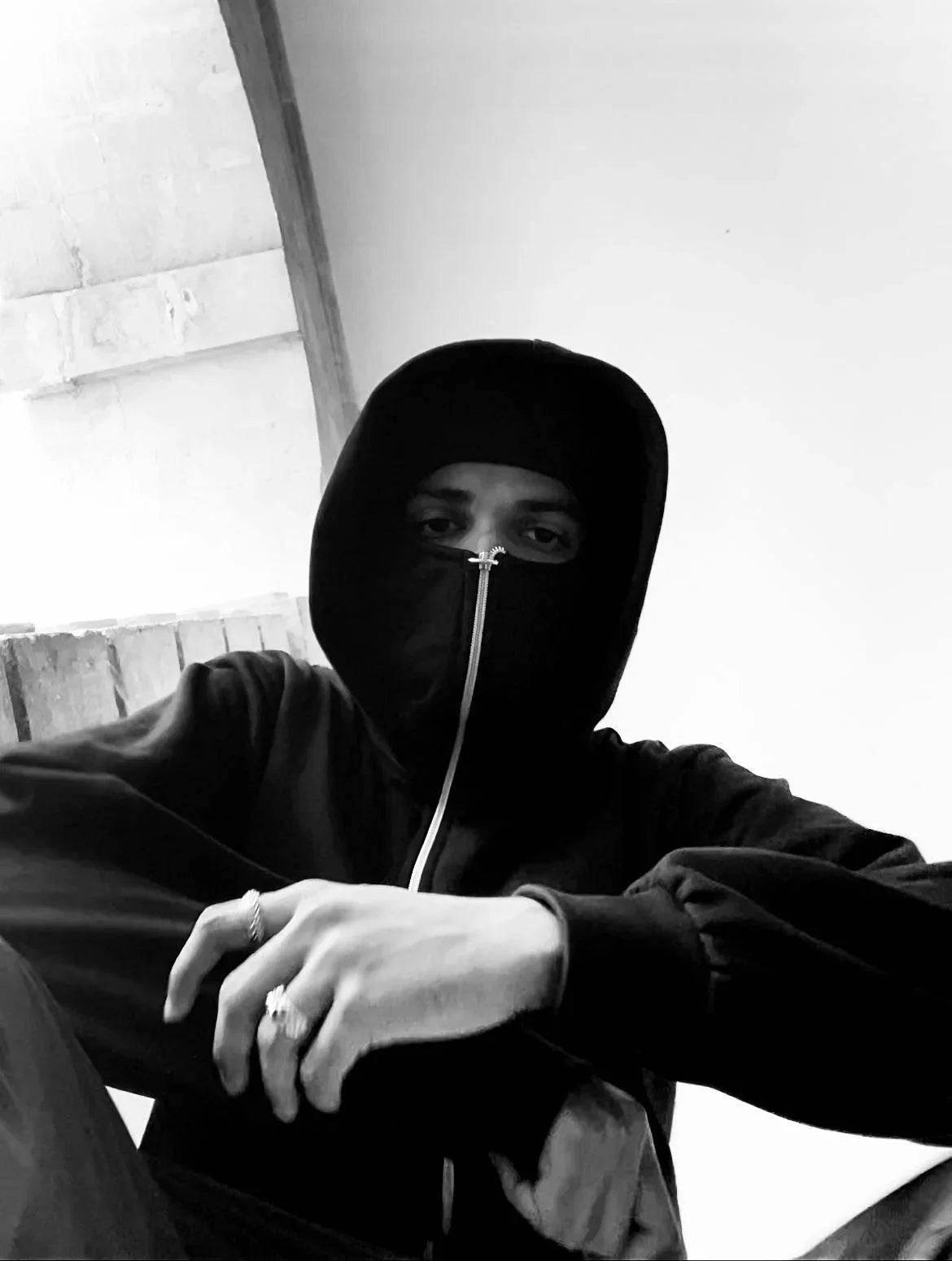 Man wearing a black zip-up balaclava hoodie and rings, seated in urban setting